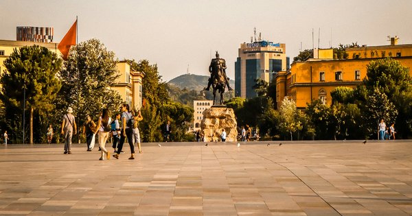 People crossing Skanderbeg Square in Tirana, Albania, with the Skanderbeg statue and Dajti mountain behind