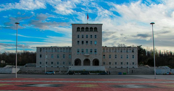 A pale stone university building with an arched entrance and a central tower, seen across a patterned plaza in Tirana under a bright blue sky