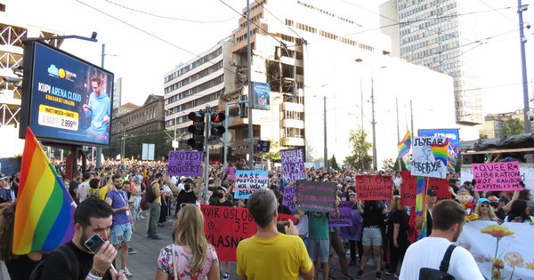 A large crowd carrying rainbow flags marches down a city street in Belgrade