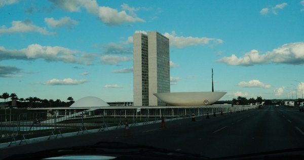 The tower and bowl-shaped chambers of Brazil's National Congress in Brasília, seen from the road under a blue sky with clouds