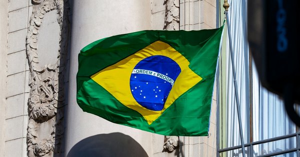 A Brazilian flag, green with a yellow diamond and a blue starry globe reading “Ordem e Progresso”, flying against a sunlit stone building