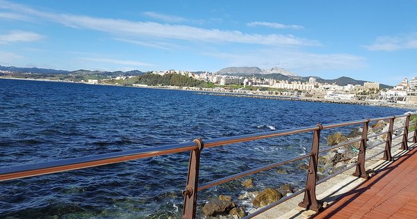 Ceuta's seafront promenade and apartment blocks curving around the bay, with mountains rising behind under a blue sky