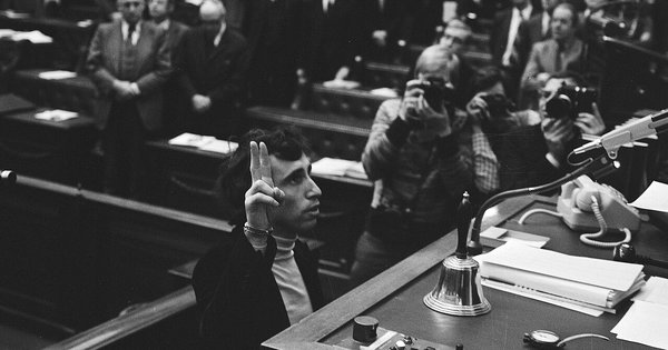 Black-and-white photo of a young Coos Huijsen raising two fingers to take the oath in the Dutch House of Representatives on 30 March 1976, as press photographers crowd around and members stand behind him