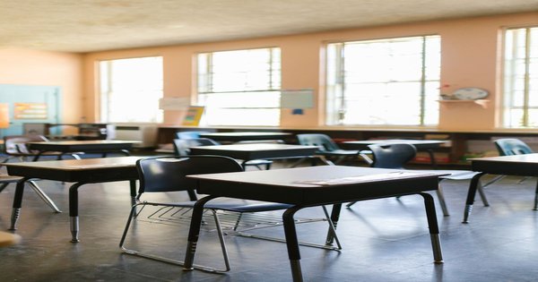 An empty classroom with rows of wooden desks and large windows letting in daylight