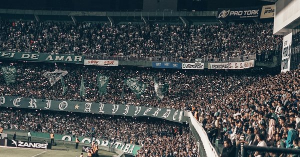 A packed football stadium at night, fans in the stands holding up scarves under the floodlights