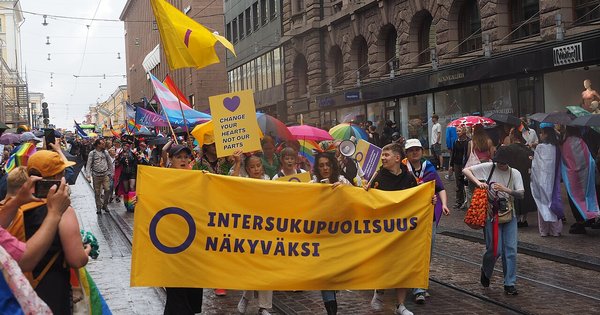 Marchers in the rain carry a yellow banner with a purple circle reading “Intersukupuolisuus näkyväksi” and a sign saying “Change your hearts not our parts” at Helsinki Pride 2023