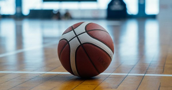 A basketball resting on the polished wooden floor of an indoor gym, with a hoop blurred in the background