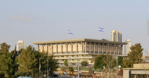The Knesset building in Jerusalem, a square stone structure with Israeli flags on its roof, seen above trees