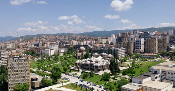 Sunny aerial view over central Pristina, Kosovo, with the domed National Library surrounded by green lawns, apartment towers and hills behind