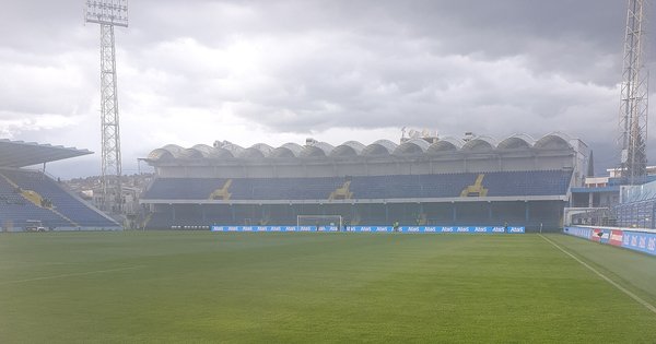 The empty pitch and blue-and-yellow stands of the Podgorica City Stadium under a grey sky