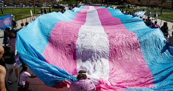 A crowd holds a huge pink, blue and white transgender pride flag stretched out across a sunny plaza