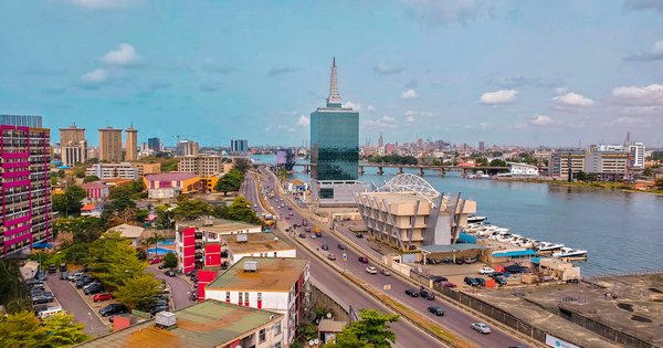 Aerial view of Lagos, Nigeria, with a glass tower, a road along the lagoon and a long bridge stretching across the water under a cloudy blue sky