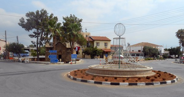 The main square of İskele (Trikomo) in northern Cyprus: a roundabout with a fountain and a wire globe sculpture, palm trees and low buildings under a pale sky