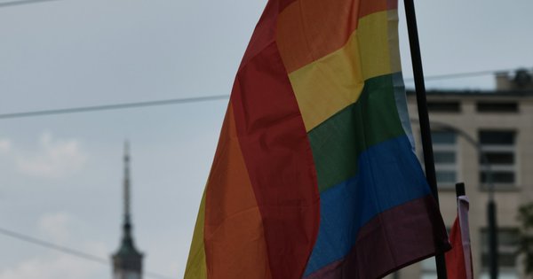 A rainbow flag flies at a Pride march in Warsaw, with the spire of the Palace of Culture and Science against a pale sky behind it