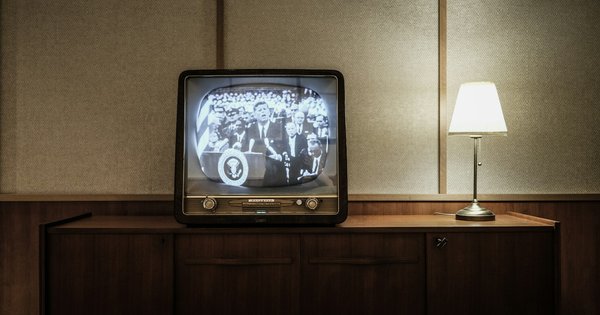 A vintage television on a wooden sideboard, beside a lit table lamp, showing a black-and-white broadcast of a politician speaking at a podium