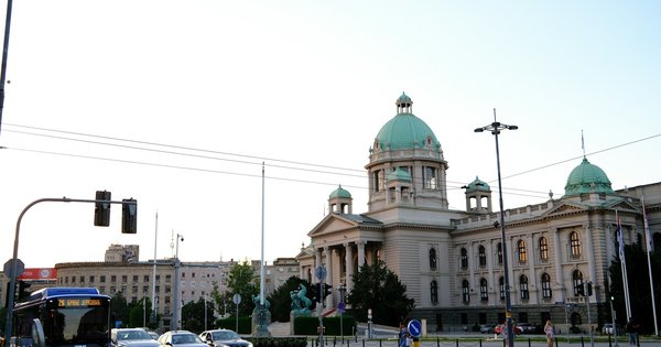 The National Assembly of Serbia in Belgrade, a cream stone building with green copper domes, seen across a busy street at dusk