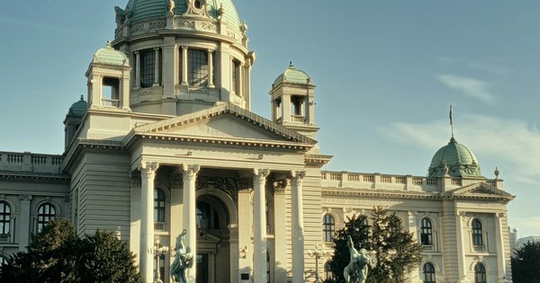 The National Assembly of Serbia in Belgrade, a cream stone building with green copper domes and bronze horse statues, under a clear blue sky