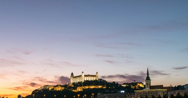 Bratislava Castle lit up at dusk above the Danube, with the city's lights reflected in the river