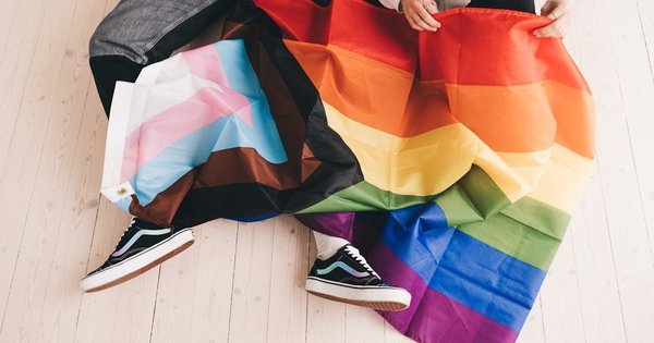 A person wrapped in a Progress Pride flag with the trans-inclusive stripes visible.