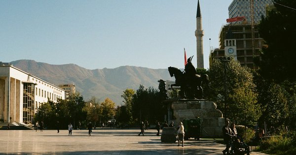 Tirana's Skanderbeg Square on a sunny afternoon, with the equestrian statue, a minaret, the clock tower and mountains behind