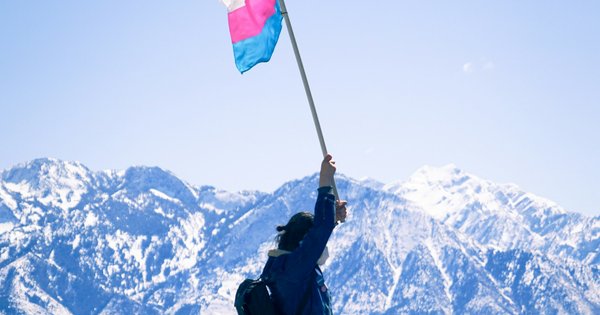A person holds a pink, white and blue transgender pride flag aloft against a backdrop of snow-covered mountains