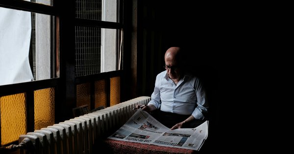 An older man in a white shirt reads a Turkish newspaper at a window table in a dim Istanbul café