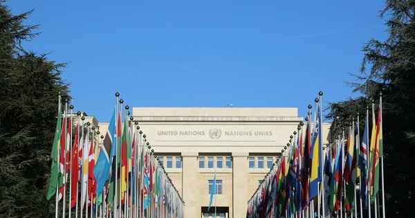Two rows of national flags lead to the United Nations building at the Palais des Nations in Geneva under a clear blue sky