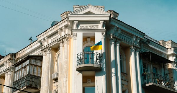 A cream neoclassical building in Kyiv with a blue and yellow Ukrainian flag hanging from its balcony under a clear sky