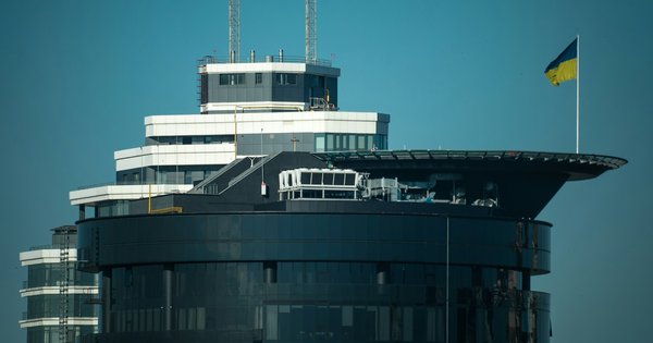 A dark glass office tower with a rooftop helipad in Kyiv, with the blue and yellow Ukrainian flag flying beside it against a clear sky